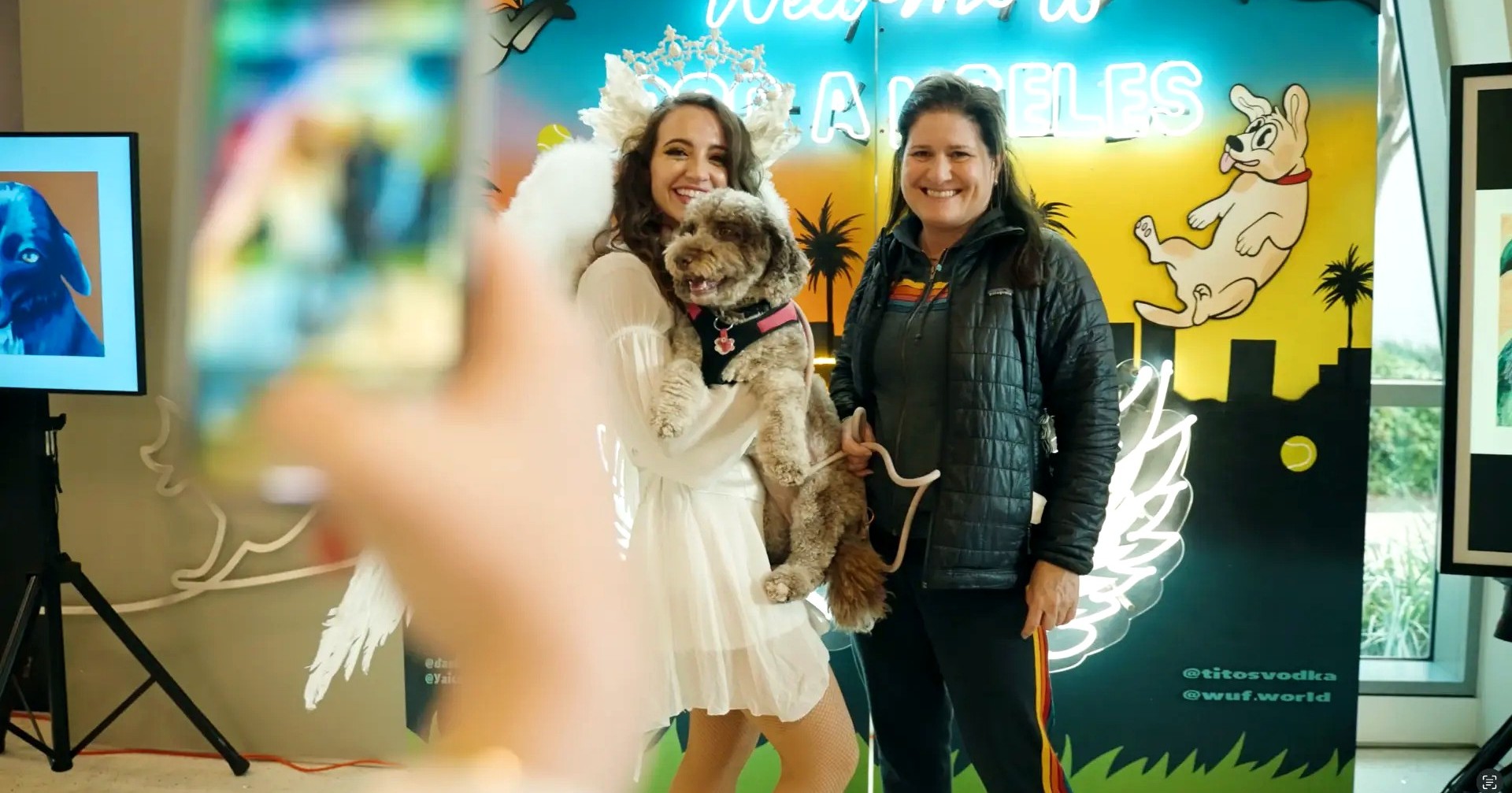 Two guests posing with a curly-coated dog in front of a Welcome to Dog Angeles neon mural
